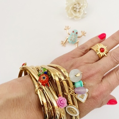 Pulseiras e anéis dourados com pendentes coloridos numa mão com unhas vermelhas, sobre fundo branco com broches de flor branca e rã azul