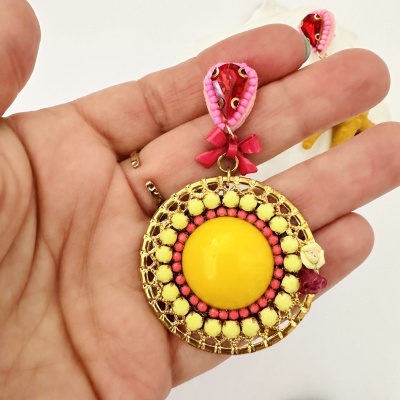 Brincos coloridos com base redonda dourada e detalhes amarelos, cor-de-rosa e vermelho, segurados numa mão.