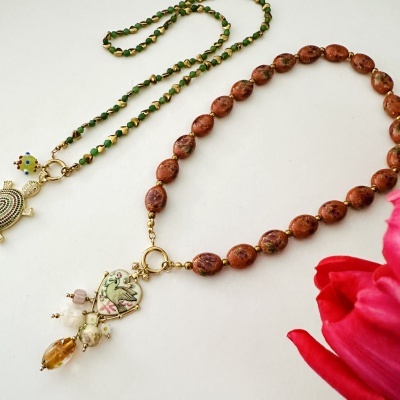 Dois colares coloridos em fundo branco com pendentes distintos e uma flor vermelha ao lado