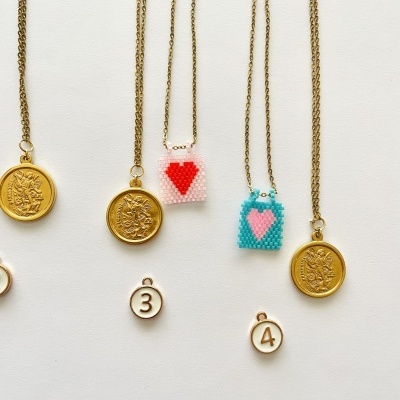 Colares com medalhões dourados e pendentes de miçangas coloridas com corações, sobre fundo branco com etiquetas numeradas.