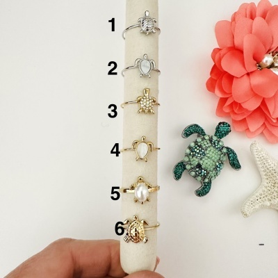 Seis anéis com tartarugas em prata e dourado, decorados com pérolas e pedras, exibidos em suporte branco com flor cor de coral, tartaruga verde brilhante e estrela do mar ao fundo.