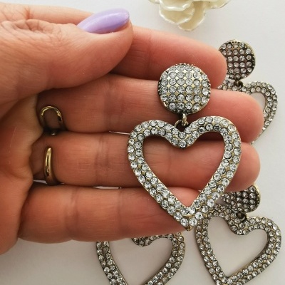 Brincos pendentes de coração com cristais brilhantes e fundo branco com flor decorativa.