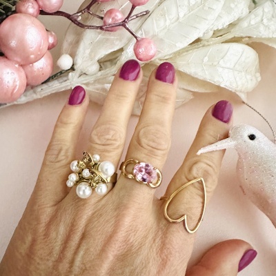 Mão com três anéis dourados e unhas roxas, sobre fundo decorativo rosa com folhas e pássaro branco.