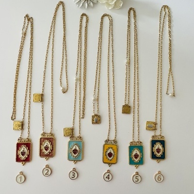 Seis colares dourados com pendentes coloridos e números, dispostos em fundo branco com flores decorativas acima