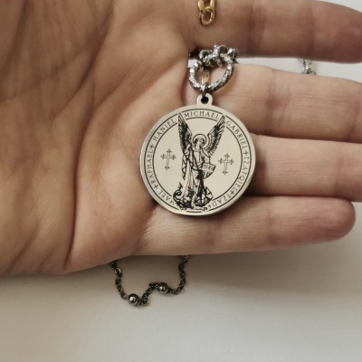 Colar com pendente redondo de metal prateado com imagem gravada de anjo e texto em volta.