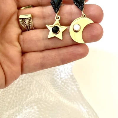 Brincos dourados de estrela e lua com pedras pretas e pérola na mão.