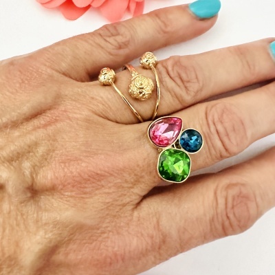 Anel dourado com esferas e pedras azuis, rosas e verdes numa mão com unhas azuis