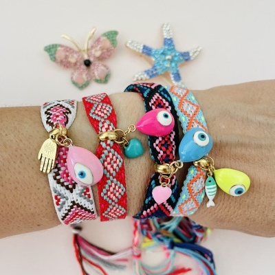 Pulseiras coloridas trançadas com pendentes de olho, coração, peixe e mão num pulso com fundo branco e decoração colorida