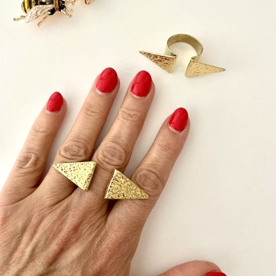 Anel dourado triangular texturizado na mão com unhas vermelhas, anel igual e brincos de abelha na superfície branca