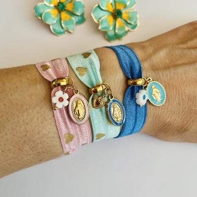 Três pulseiras de tecido com corações dourados e pendentes flor e medalha dourada usadas no pulso, com brincos florais verdes e amarelos ao fundo.