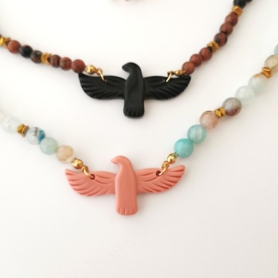 Dois colares com pendentes de pássaros, um preto e outro salmão, com contas coloridas em fundo branco.