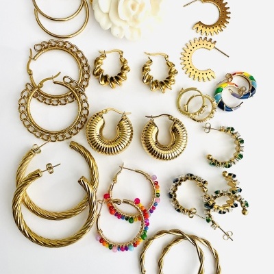 Vários pares de brincos dourados com diferentes designs e cores sobre fundo branco.