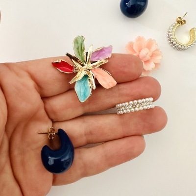 Conjunto de brincos coloridos e dourados sobre fundo branco, com um brinco flor em esmalte nas cores vermelho, verde, lilás, azul e pêssego, brincos meia-lua azul escuro e brinco dourado com pérolas.
