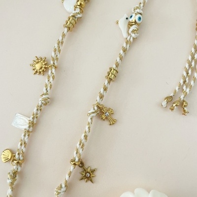 Acessório trançado branco e dourado com pendentes dourados de várias formas sobre fundo bege.