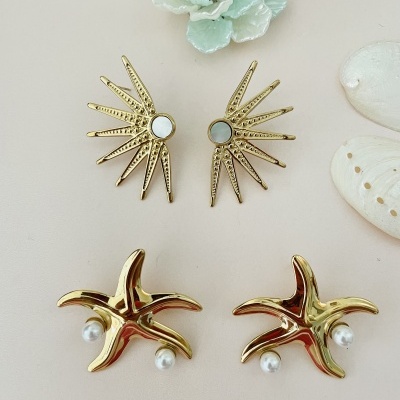 Dois pares de brincos dourados, um estrela do mar com pérolas e outro com raios espelhados, sobre fundo claro com flor e conchas