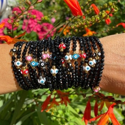 Pulseiras com contas pretas e contas coloridas de olho grego no braço de uma pessoa, em ambiente exterior com flores.