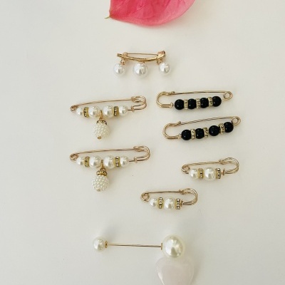 Alfinetes de segurança decorativos dourados com pérolas e contas pretas sobre superfície branca com flor rosa