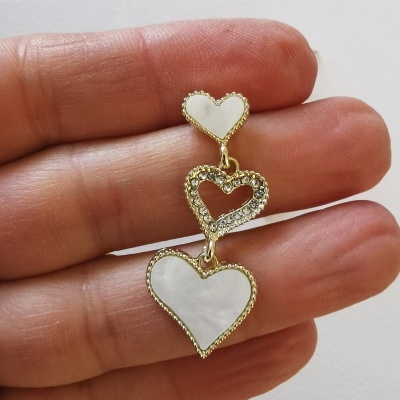 Brinco dourado com três corações pendurados, detalhes brancos perolados e vazados, segurado por mão