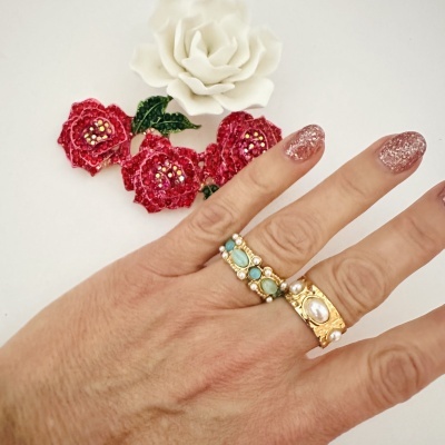 Mão com anéis dourados e pérolas, unhas rosa brilhante, flores artificiais brancas e vermelhas ao fundo