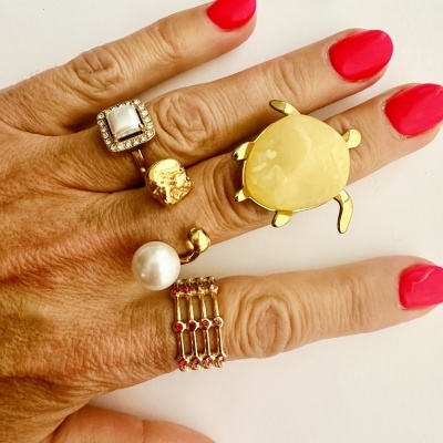 Mão feminina com cinco anéis dourados, unhas vermelhas, fundo branco.