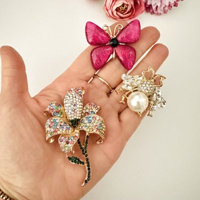Três pins decorativos de borboleta, flor e abelha em mão, fundo branco com flores desfocadas