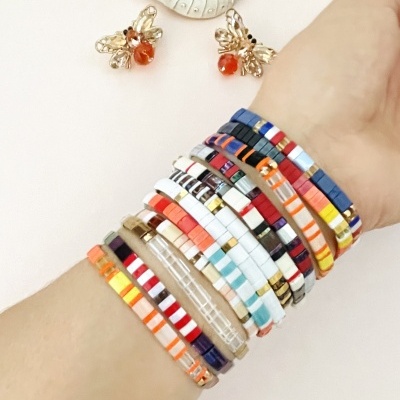 Pulseiras coloridas de contas quadradas no pulso com brincos dourados em forma de abelha e concha branca ao fundo.