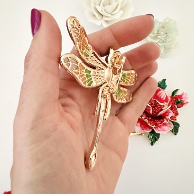 Broche dourado em forma de libélula com detalhes verdes, segurado por mão, fundo branco com flores decorativas