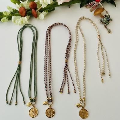 Três colares com medalhões dourados e cordões coloridos sobre superfície branca com flores artificiais e broche de libélula