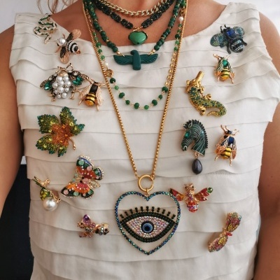 Colares verdes com pendente de coração e broches coloridos em blusa branca em camadas.