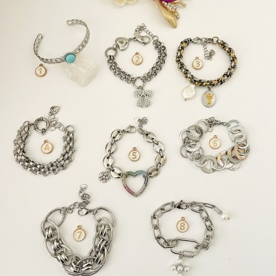 Oito pulseiras metálicas numeradas dispostas em fundo branco, incluindo uma dourada com flor branca e pedra roxa.