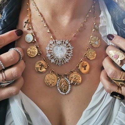 Colar dourado com pendentes de moedas usado por pessoa com casaco preto e blusa branca, com vários anéis nas mãos