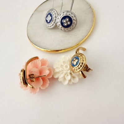Brincos com flores decorativas em coro dourada e azul, apresentados em suporte branco com borda dourada