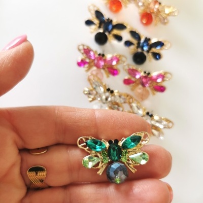 Anel em forma de borboleta com pedras coloridas, segurado por mão, vários anéis similares ao fundo