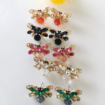 Cinco pares de brincos abelha com pedras coloridas em fundo branco e flores amarelas