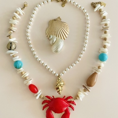 Três colares com conchas, pérolas e pendentes coloridos sobre fundo bege.