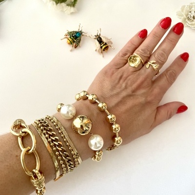 Pulso com pulseiras douradas e pérolas, anéis dourados, unhas vermelhas e brincos de abelha ao fundo