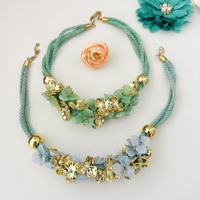 Conjunto de colar e pulseira com flores douradas e coloridas sobre fundo branco com flores artificiais.