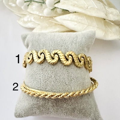 Duas pulseiras douradas em suporte cinzento com folhas decorativas atrás