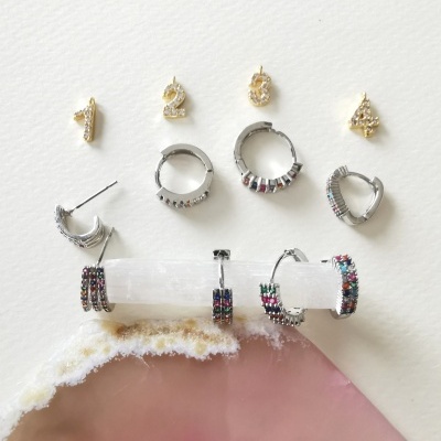 Conjunto de brincos dourados e prateados com números e pedras coloridas sobre fundo branco e rosa marmoreado.