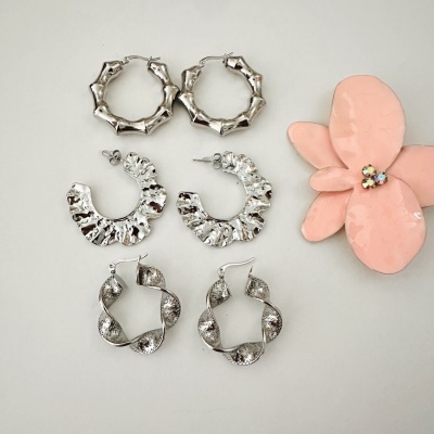 Três pares de brincos prateados com texturas variadas e uma flor rosa decorativa ao lado