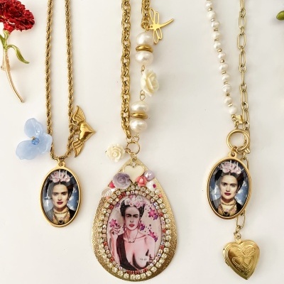 Três colares com pendentes com imagens de Frida Kahlo e acessórios dourados, apresentados sobre fundo branco com flores decorativas.