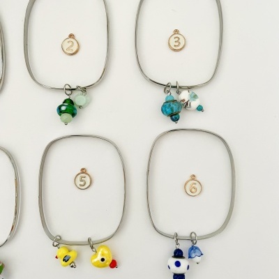 Braceletes quadrados metálicos com pendentes coloridos e etiquetas numéricas em fundo branco.