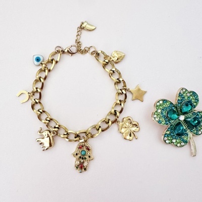 Pulseira dourada com vários pendentes e broche trevo azul com pedras.