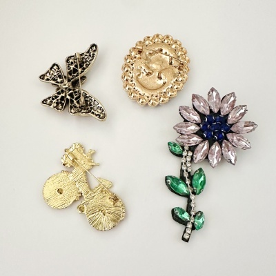 Broches em metal vistos de trás, incluindo borboleta, elipse, bicicleta e flor colorida.