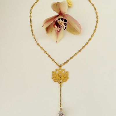 Colar dourado com pingente de flor de lótus e cristal, ao lado de flor natural rosa e creme.