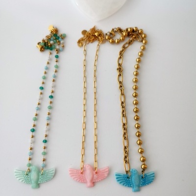 Três colares com pendentes de pássaros em verde, rosa e azul com diferentes tipos de correntes douradas sobre fundo branco.