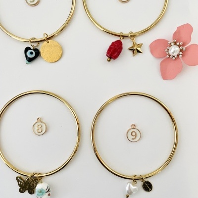 Quatro pulseiras douradas com pendentes variados sobre fundo branco e flor rosa