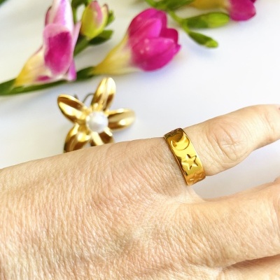 Anel dourado com estrelas recortadas num dedo, anel de flor dourada com pérola, flores coloridas ao fundo