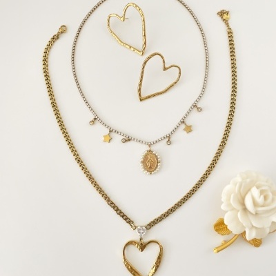 Conjunto de bijuteria com colares e brincos em forma de coração e corrente com pingentes, sobre fundo branco com flor decorativa.