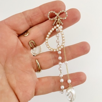 Brinco de pérolas com corrente dourada e laço, sobre uma mão com anéis